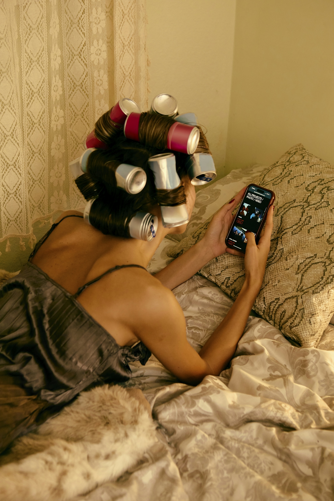 A woman lying on a bed with vintage curlers in her hair, looking at her phone in warm afternoon light